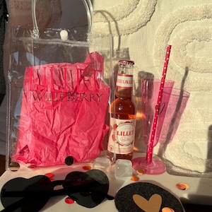 K&ouml;nnte beinhalten: Eine transparente Tragetasche mit Griff, gef&uuml;llt mit rosa Seidenpapier mit der Aufschrift "LILLET WILD BERRY". Eine Flasche Lillet Berry, herzf&ouml;rmige Sonnenbrille, ein rosa Strohhalm und ein Untersetzer mit Herzdesign sind ebenfalls vorhanden.