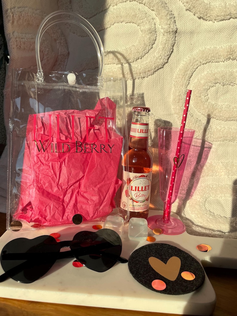 K&ouml;nnte beinhalten: Eine transparente Tragetasche mit Griff, gef&uuml;llt mit rosa Seidenpapier mit der Aufschrift "LILLET WILD BERRY". Eine Flasche Lillet Berry, herzf&ouml;rmige Sonnenbrille, ein rosa Strohhalm und ein Untersetzer mit Herzdesign sind ebenfalls vorhanden.