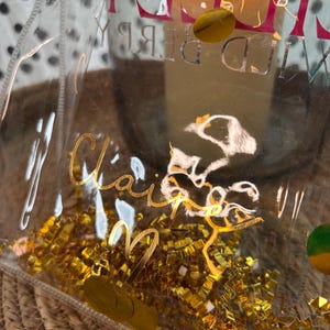 K&ouml;nnte beinhalten: Eine transparente Plastikt&uuml;te mit goldenen Buchstaben, die "Claire" und "Wild Berry" zeigen. Die T&uuml;te ist mit goldenem Metallic-Konfetti gef&uuml;llt und liegt auf einer gewebten Strohoberfl&auml;che. Der Hintergrund ist unscharf.