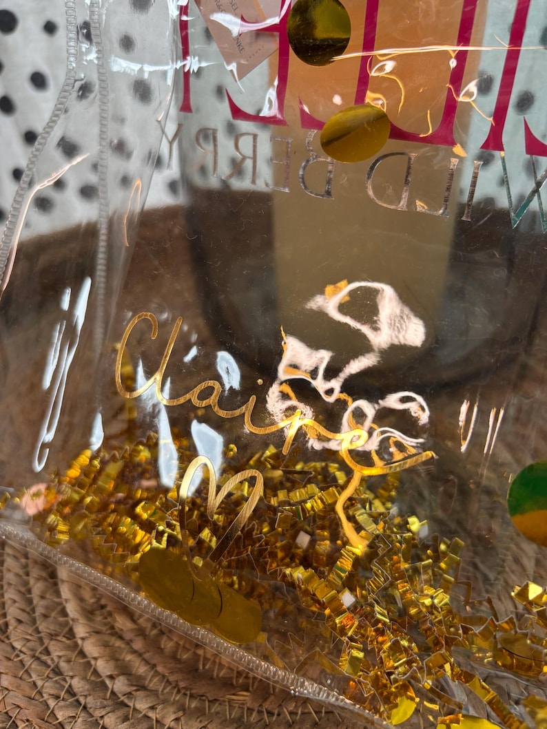 K&ouml;nnte beinhalten: Eine transparente Plastikt&uuml;te mit goldenen Buchstaben, die "Claire" und "Wild Berry" zeigen. Die T&uuml;te ist mit goldenem Metallic-Konfetti gef&uuml;llt und liegt auf einer gewebten Strohoberfl&auml;che. Der Hintergrund ist unscharf.