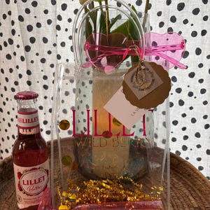 K&ouml;nnte beinhalten: Eine transparente Tragetasche mit dem Text "Lillet Wild Berry" enth&auml;lt eine Flasche Lillet Berry, eine rosa herzf&ouml;rmige Sonnenbrille und ein rosa Sektglas. Die Tasche enth&auml;lt auch goldenes Lametta und eine Vase mit wei&szlig;en Tulpen.