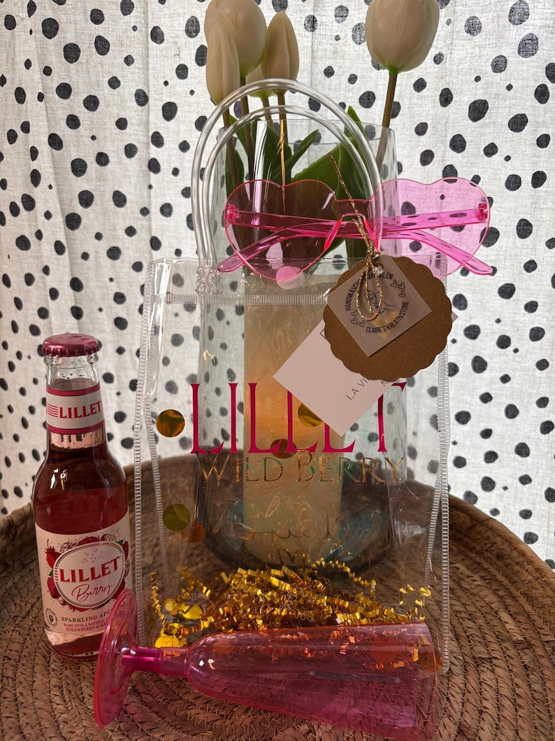 K&ouml;nnte beinhalten: Eine transparente Tragetasche mit dem Text "Lillet Wild Berry" enth&auml;lt eine Flasche Lillet Berry, eine rosa herzf&ouml;rmige Sonnenbrille und ein rosa Sektglas. Die Tasche enth&auml;lt auch goldenes Lametta und eine Vase mit wei&szlig;en Tulpen.