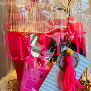 May include: A clear cellophane gift bag filled with pink and red items. The bag contains a small bottle, a pink heart-shaped bottle, and a tag with text. The bag has the word "LIEBE" in large pink letters.