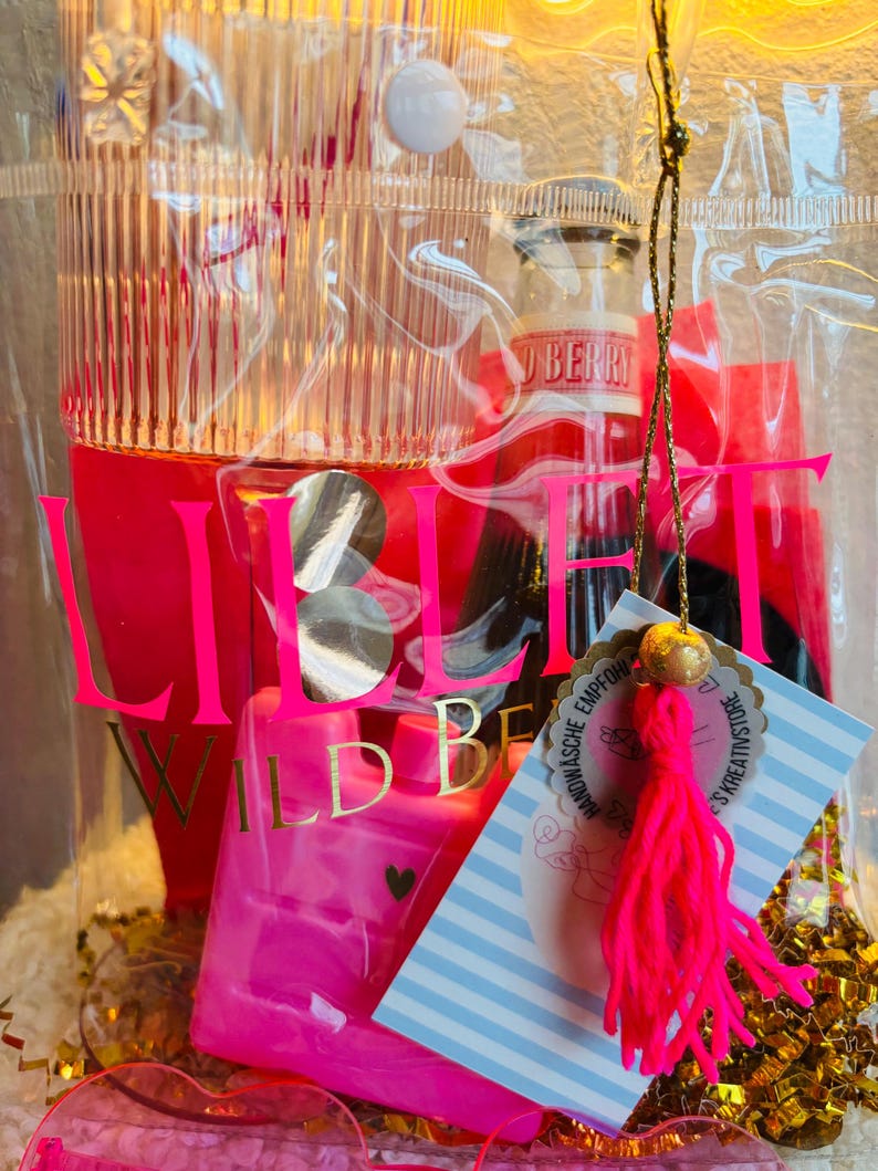 May include: A clear cellophane gift bag filled with pink and red items. The bag contains a small bottle, a pink heart-shaped bottle, and a tag with text. The bag has the word "LIEBE" in large pink letters.