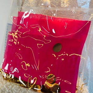 May include: A clear plastic tote bag with a hot pink envelope inside. The envelope has gold accents and a gold heart charm. The bag is filled with gold confetti and small, round, brown objects.