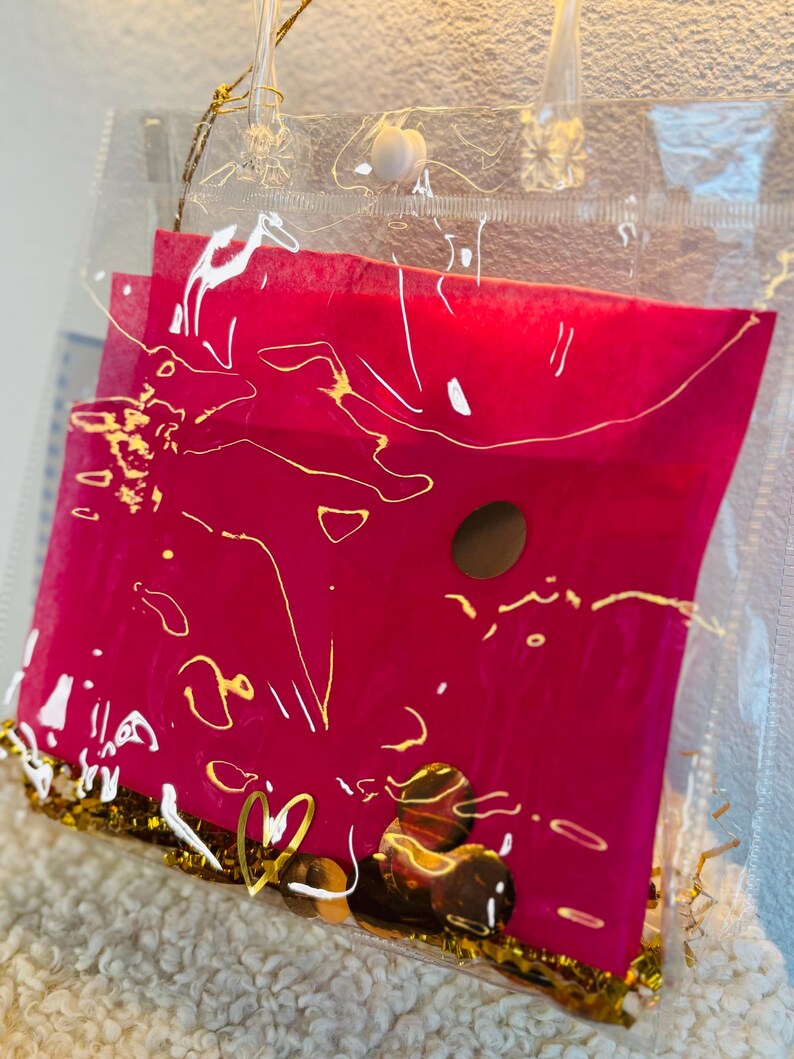 May include: A clear plastic tote bag with a hot pink envelope inside. The envelope has gold accents and a gold heart charm. The bag is filled with gold confetti and small, round, brown objects.