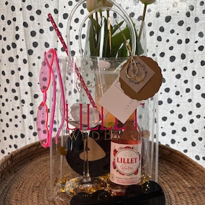 K&ouml;nnte beinhalten: Eine transparente Tragetasche mit einer Flasche Lillet Berry, einem Weinglas und herzf&ouml;rmigen Sonnenbrillen. Die Tasche enth&auml;lt auch eine rosa Sonnenbrille und wei&szlig;e Tulpen. Auf der Tragetasche steht "Lillet Wild Berry".