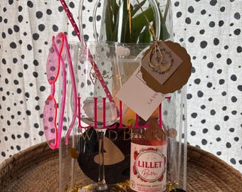 May include: A clear tote bag filled with a bottle of Lillet Berry sparkling aperitif, a wine glass, and a pair of heart-shaped sunglasses. The bag also contains a pair of pink sunglasses and a bouquet of white tulips. The tote bag has the words "Lillet Wild Berry" printed on it.