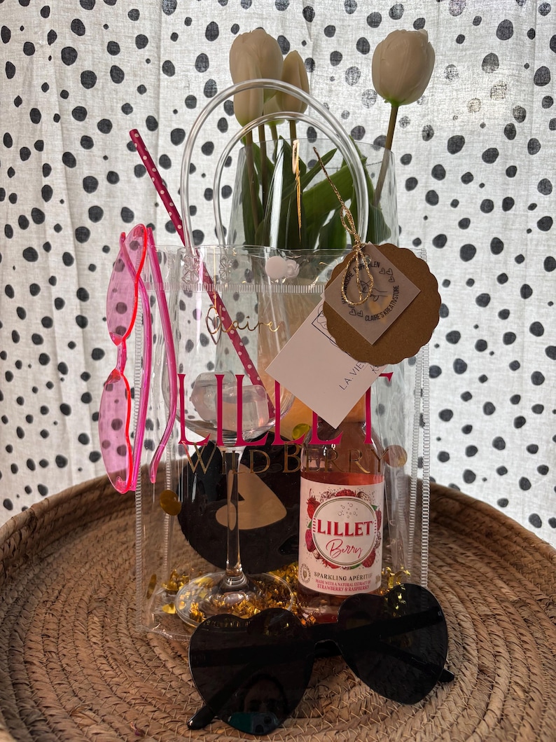 K&ouml;nnte beinhalten: Eine transparente Tragetasche mit einer Flasche Lillet Berry, einem Weinglas und herzf&ouml;rmigen Sonnenbrillen. Die Tasche enth&auml;lt auch eine rosa Sonnenbrille und wei&szlig;e Tulpen. Auf der Tragetasche steht "Lillet Wild Berry".