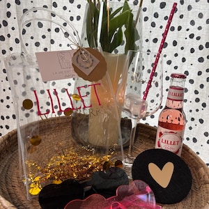 K&ouml;nnte beinhalten: Eine transparente Tragetasche mit dem Text "Lillet Berry" und goldenem Konfetti, eine Flasche Lillet Berry, herzf&ouml;rmige Sonnenbrillen und ein schwarzer Untersetzer mit goldenem Herz. Wei&szlig;e Tulpen befinden sich in einer Glasvase.