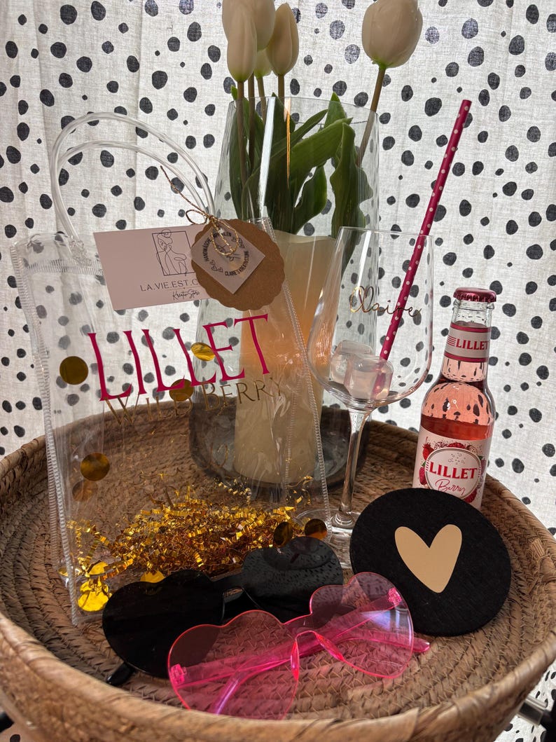 K&ouml;nnte beinhalten: Eine transparente Tragetasche mit dem Text "Lillet Berry" und goldenem Konfetti, eine Flasche Lillet Berry, herzf&ouml;rmige Sonnenbrillen und ein schwarzer Untersetzer mit goldenem Herz. Wei&szlig;e Tulpen befinden sich in einer Glasvase.
