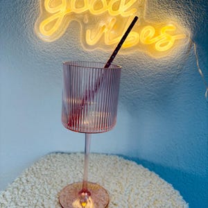 May include: A pink, ribbed-glass goblet with a long stem and round base. A black and red striped straw is inside the glass. The words "good vibes" are written in yellow neon in the background.