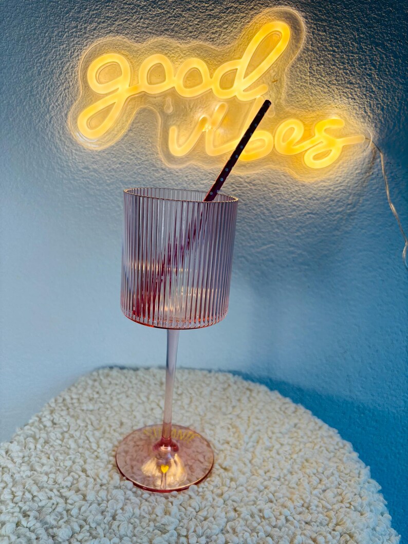 May include: A pink, ribbed-glass goblet with a long stem and round base. A black and red striped straw is inside the glass. The words "good vibes" are written in yellow neon in the background.