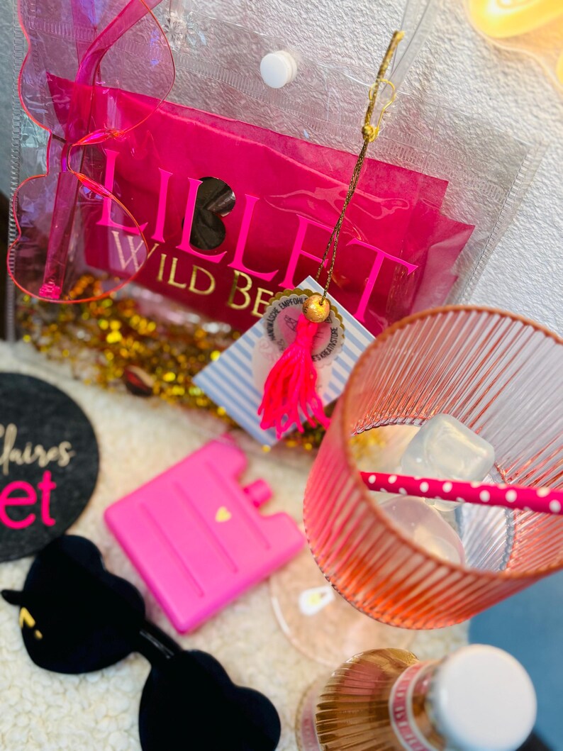 May include: A clear tote bag with a pink package that reads "LILLET WILD BERRY". The image also shows a pink ice pack, a pink and white striped glass, and a black heart-shaped object.