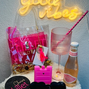 May include: A neon sign that reads "good vibes" illuminates the scene. A clear tote bag contains pink items, including sunglasses, a drink glass, and a bottle of Wild Berry beverage. A coaster with "Claire's Lillet" is also visible.
