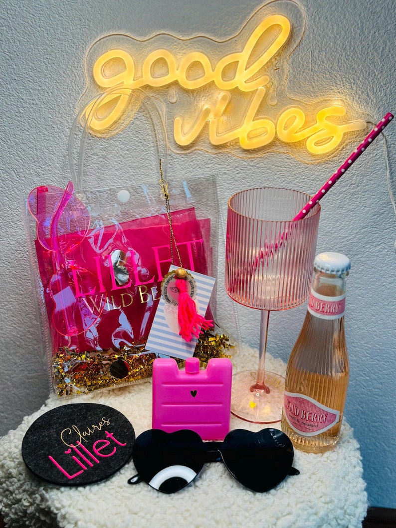 May include: A neon sign that reads "good vibes" illuminates the scene. A clear tote bag contains pink items, including sunglasses, a drink glass, and a bottle of Wild Berry beverage. A coaster with "Claire's Lillet" is also visible.