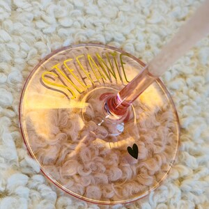 May include: A pink-tinted wine glass with the name "STEFANIE" in yellow script. A small black heart is near the edge of the glass. The glass is set against a white, fluffy background.