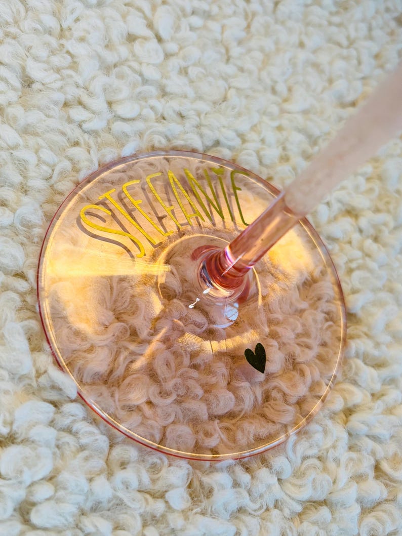 May include: A pink-tinted wine glass with the name "STEFANIE" in yellow script. A small black heart is near the edge of the glass. The glass is set against a white, fluffy background.