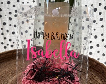 May include: A clear plastic tote bag with a handle, filled with pink shredded paper and black filler. The bag has the words "HAPPY BIRTHDAY Isabella" in pink lettering.