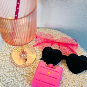 May include: A pink, ribbed glass with a polka-dot straw, heart-shaped sunglasses, and a pink ice pack. The sunglasses have black and pink lenses. The ice pack has a gold heart detail. The items are on a white, textured surface.