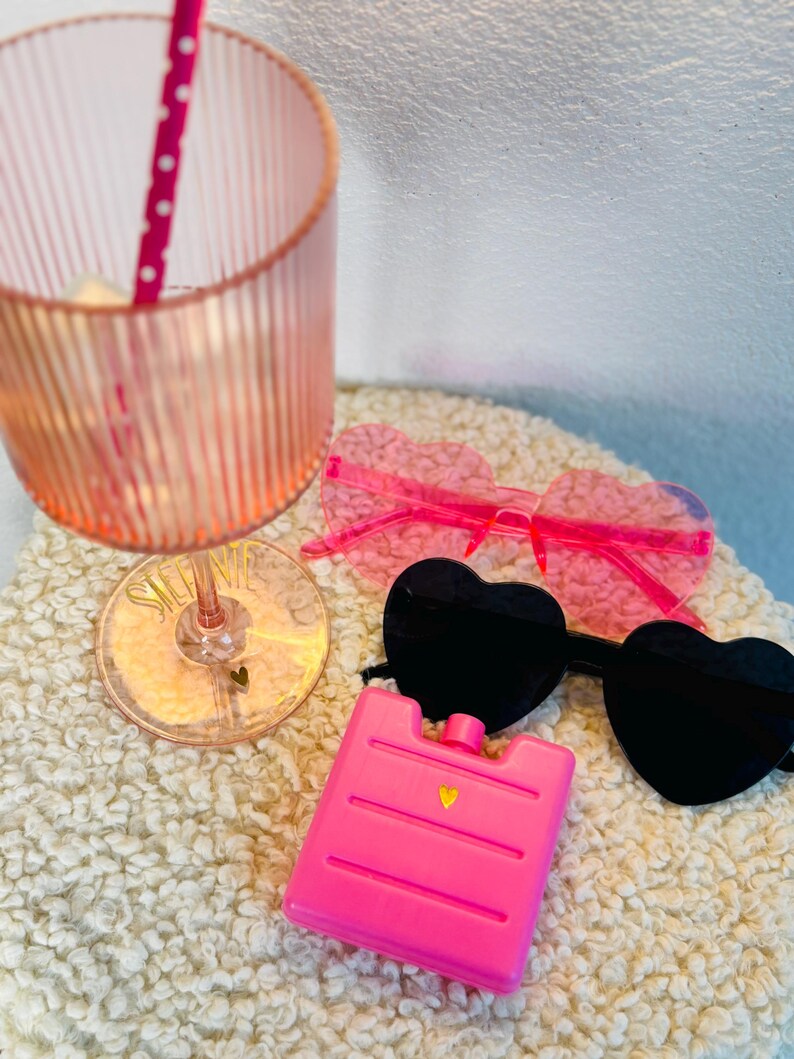 May include: A pink, ribbed glass with a polka-dot straw, heart-shaped sunglasses, and a pink ice pack. The sunglasses have black and pink lenses. The ice pack has a gold heart detail. The items are on a white, textured surface.