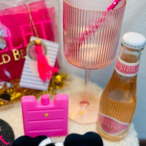 May include: A collection of pink accessories, including a ribbed glass, a bottle of Wild Berry soda, a pink ice pack, and heart-shaped sunglasses. A pink and white polka dot straw is in the glass. A gift bag and a tag with a tassel are in the background.