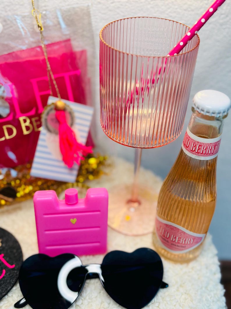 May include: A collection of pink accessories, including a ribbed glass, a bottle of Wild Berry soda, a pink ice pack, and heart-shaped sunglasses. A pink and white polka dot straw is in the glass. A gift bag and a tag with a tassel are in the background.