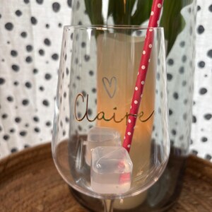 K&ouml;nnte beinhalten: Ein klares Weinglas mit dem Namen "Claire" in goldener Schrift und einem kleinen Herz. Das Glas enth&auml;lt Eisw&uuml;rfel und einen rot-wei&szlig; gepunkteten Strohhalm. Das Glas steht auf einem Holztablett.