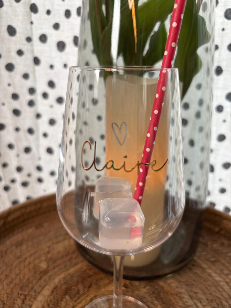 K&ouml;nnte beinhalten: Ein klares Weinglas mit dem Namen "Claire" in goldener Schrift und einem kleinen Herz. Das Glas enth&auml;lt Eisw&uuml;rfel und einen rot-wei&szlig; gepunkteten Strohhalm. Das Glas steht auf einem Holztablett.