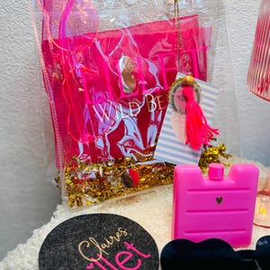 May include: A clear plastic bag with the words "WILD BE" in pink, filled with pink items and gold confetti. A pink ice pack, a black heart-shaped sunglasses, and a gray coaster with the words "Claire's Lillet" are also visible.
