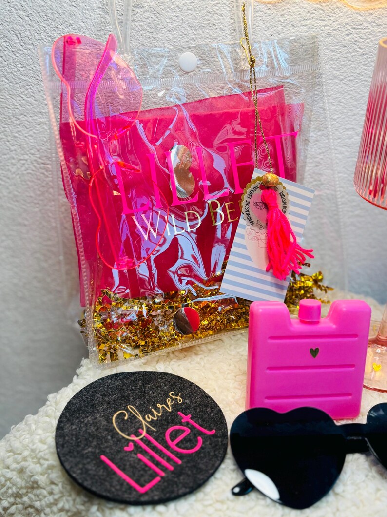 May include: A clear plastic bag with the words "WILD BE" in pink, filled with pink items and gold confetti. A pink ice pack, a black heart-shaped sunglasses, and a gray coaster with the words "Claire's Lillet" are also visible.