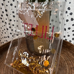 K&ouml;nnte beinhalten: Eine transparente Plastiktasche mit goldenem Konfetti und goldenen Folienakzenten. Die Tasche hat einen rosa-goldenen Text mit der Aufschrift "IT'S THE LITTLE THINGS" und "Claire M". Die Tasche steht auf einem geflochtenen Korb.