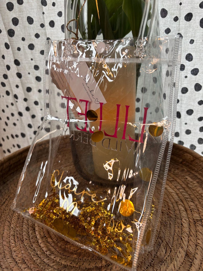 K&ouml;nnte beinhalten: Eine transparente Plastiktasche mit goldenem Konfetti und goldenen Folienakzenten. Die Tasche hat einen rosa-goldenen Text mit der Aufschrift "IT'S THE LITTLE THINGS" und "Claire M". Die Tasche steht auf einem geflochtenen Korb.