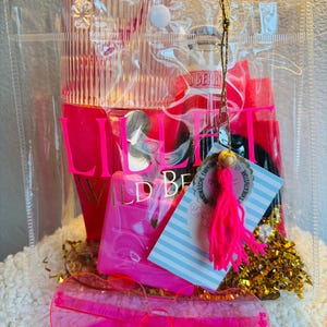 May include: A clear tote bag filled with pink items, including a tumbler, sunglasses, and a perfume bottle. The bag also contains a tag with text and a bright pink tassel. Gold confetti adds a festive touch.