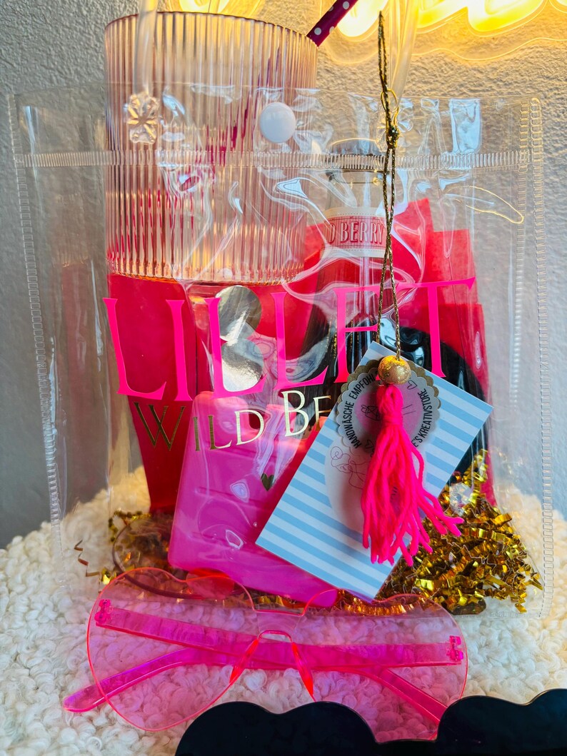 May include: A clear tote bag filled with pink items, including a tumbler, sunglasses, and a perfume bottle. The bag also contains a tag with text and a bright pink tassel. Gold confetti adds a festive touch.