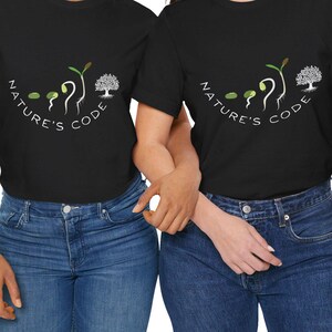 Puede incluir: Dos camisetas negras con el texto "NATURE'S CODE" en arco sobre un gráfico de una semilla germinando y un árbol. Las camisetas se combinan con vaqueros azules. La imagen está sobre un fondo blanco.