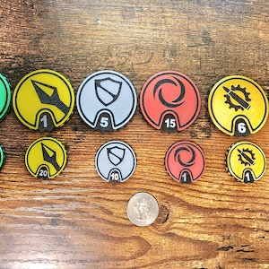 May include: A collection of twelve circular game tokens in two sizes, featuring various icons including shields, gears, and abstract symbols in green, yellow, gray, and red. Each token displays a numerical value. A US quarter coin is included for scale.