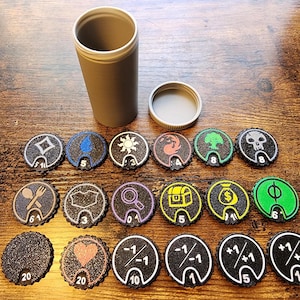 May include: A cylindrical grey storage container with a matching lid and a collection of eighteen circular game tokens. The tokens feature various icons including symbols for mana, health, treasure, and numerical modifiers with white outlines.