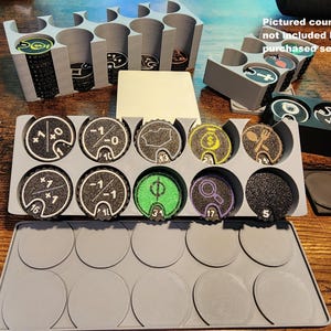 May include: A set of light gray 3D-printed plastic storage trays for circular game tokens. The trays feature multiple cylindrical slots for organizing tokens with various icons and numbers, including symbols for money, food, and magnifying glasses.