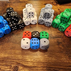 May include: A collection of six-sided dice arranged in five distinct groups on a wooden surface. The dice are colored blue, black, white, gray, green, and red, each featuring black or white circular pips indicating numerical values from one to six.