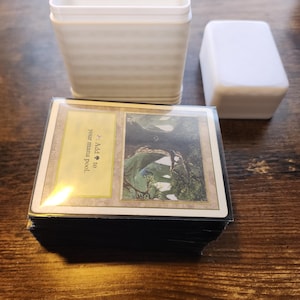 May include: A stack of trading cards with a card case and lid. The cards have a gold border and a landscape image. The text on the card reads "Add to your mana pool."