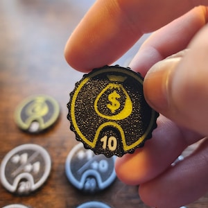 May include: A circular black game token with a scalloped edge featuring a yellow money bag icon and the number 10 printed in white. The token has a textured surface and is part of a collection of similar game pieces with various numerical values.