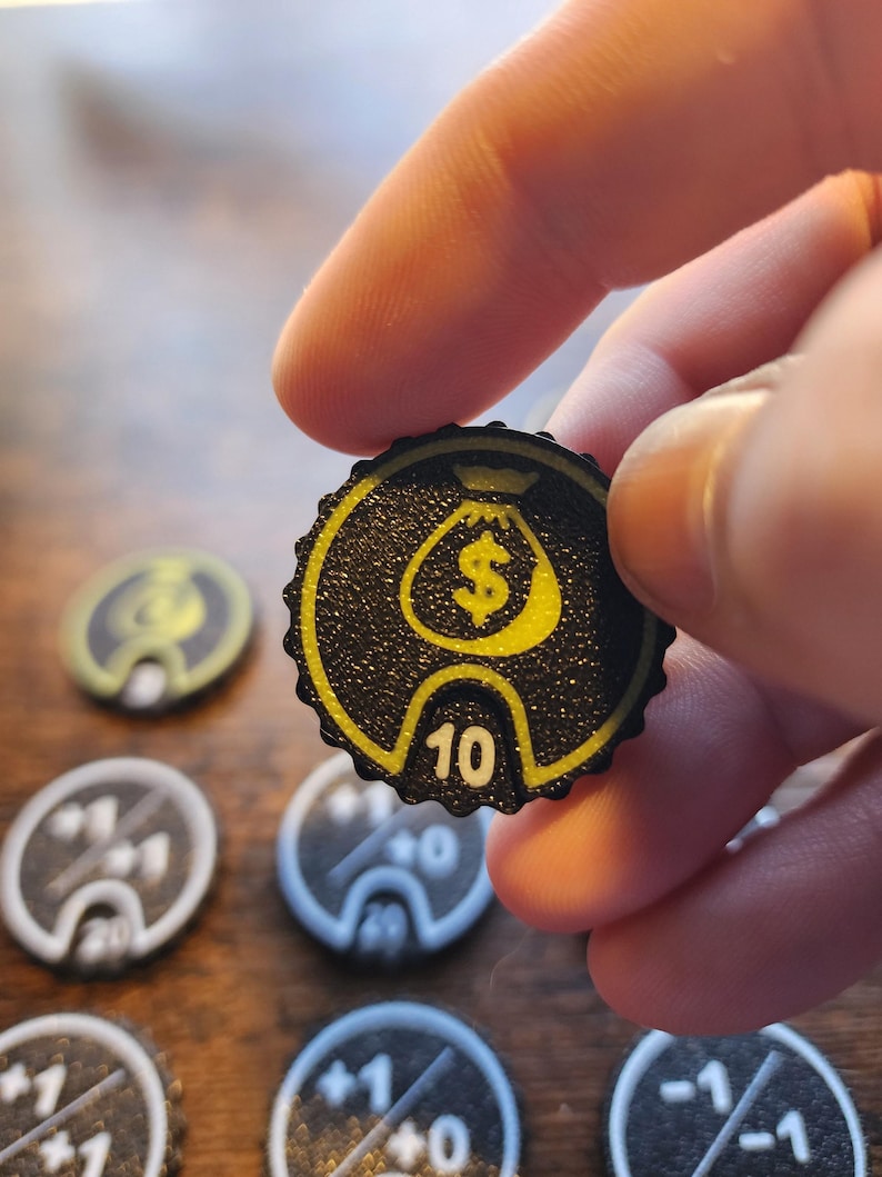 May include: A circular black game token with a scalloped edge featuring a yellow money bag icon and the number 10 printed in white. The token has a textured surface and is part of a collection of similar game pieces with various numerical values.