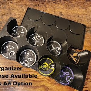 May include: A black 3D-printed plastic organizer tray containing several circular game tokens with various symbols and numbers, including plus and minus signs, a dollar sign, a spoon and fork icon, and a magnifying glass. Text reads: Organizer Case Available As An Option.