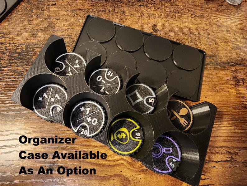 May include: A black 3D-printed plastic organizer tray containing several circular game tokens with various symbols and numbers, including plus and minus signs, a dollar sign, a spoon and fork icon, and a magnifying glass. Text reads: Organizer Case Available As An Option.