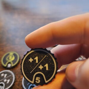 May include: A circular black game token with a scalloped edge featuring white printed text and graphics. The token displays the text +1 over +1 separated by a diagonal line, with the number 5 printed inside a white outlined arch at the bottom.