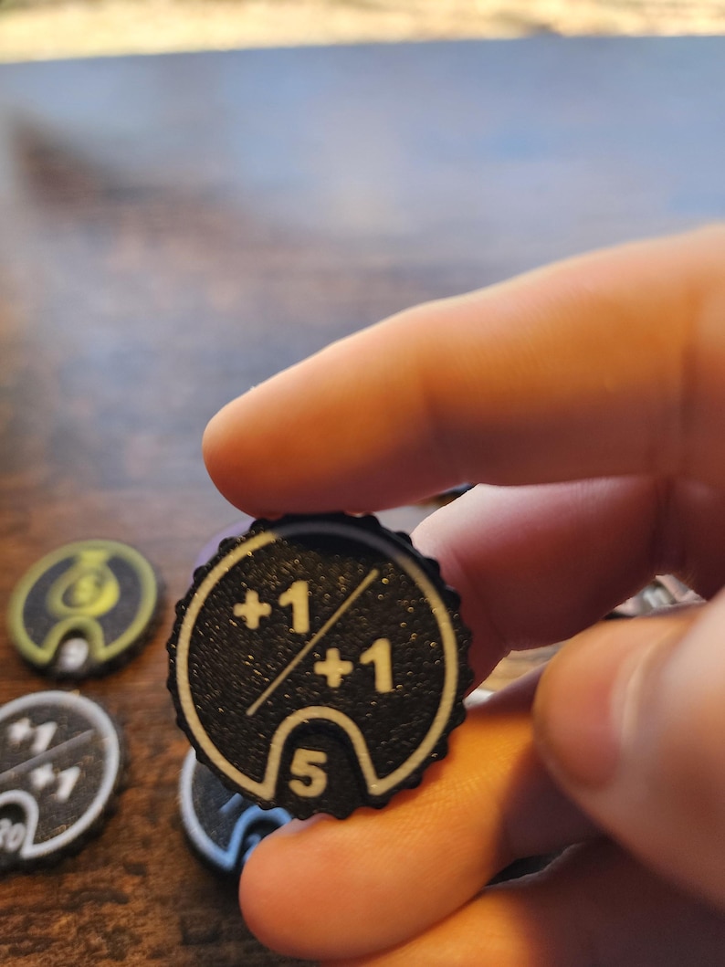 May include: A circular black game token with a scalloped edge featuring white printed text and graphics. The token displays the text +1 over +1 separated by a diagonal line, with the number 5 printed inside a white outlined arch at the bottom.