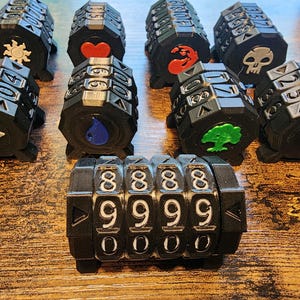 May include: A collection of black plastic gaming life counters featuring rotating number dials and various colored icons including a sun, heart, skull, water drop, and tree. Each counter has four columns of numbers and distinct symbols on the ends.