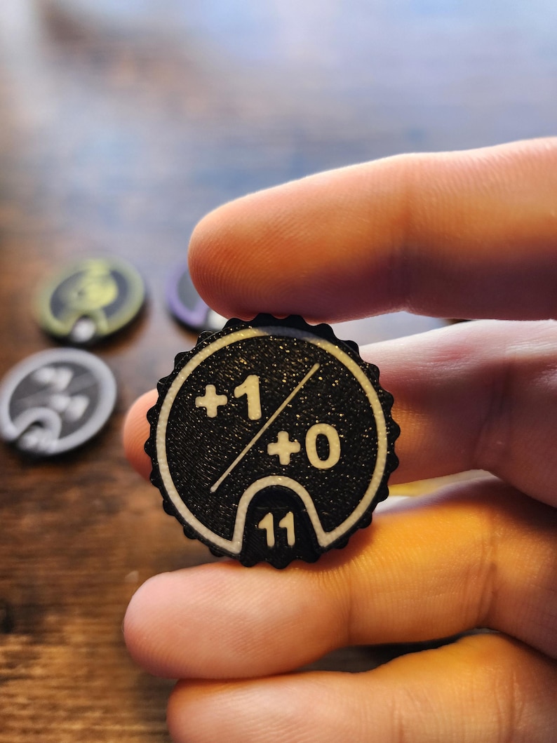 May include: A circular black game token with a scalloped edge, featuring a white printed design that includes the text +1 and +0 separated by a diagonal line, with the number 11 displayed in a small semi-circular cutout at the bottom.