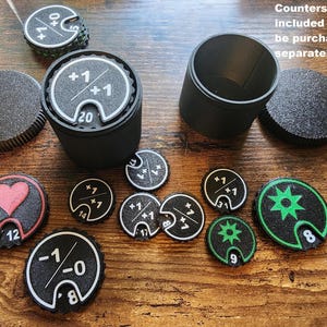 May include: A set of black circular game counters with white, red, and green printed symbols and numbers, including plus and minus signs, hearts, and star icons. Two black cylindrical containers with matching lids are included for storage.